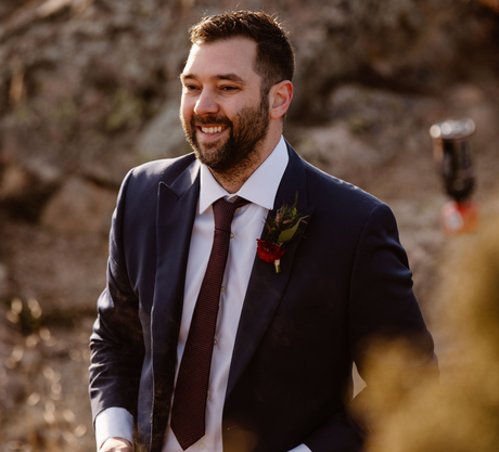 Man in a blue suit smiles outside; burgundy tie, boutonniere.