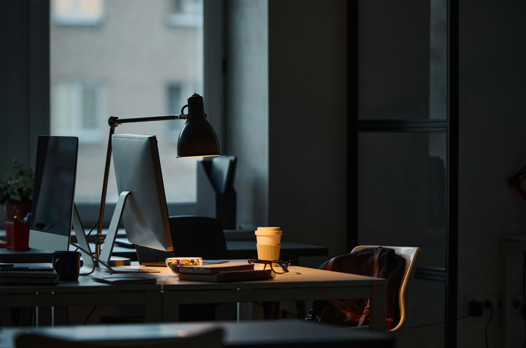 Desk in a dimly lit office, with computers, a lamp, and a coffee cup.