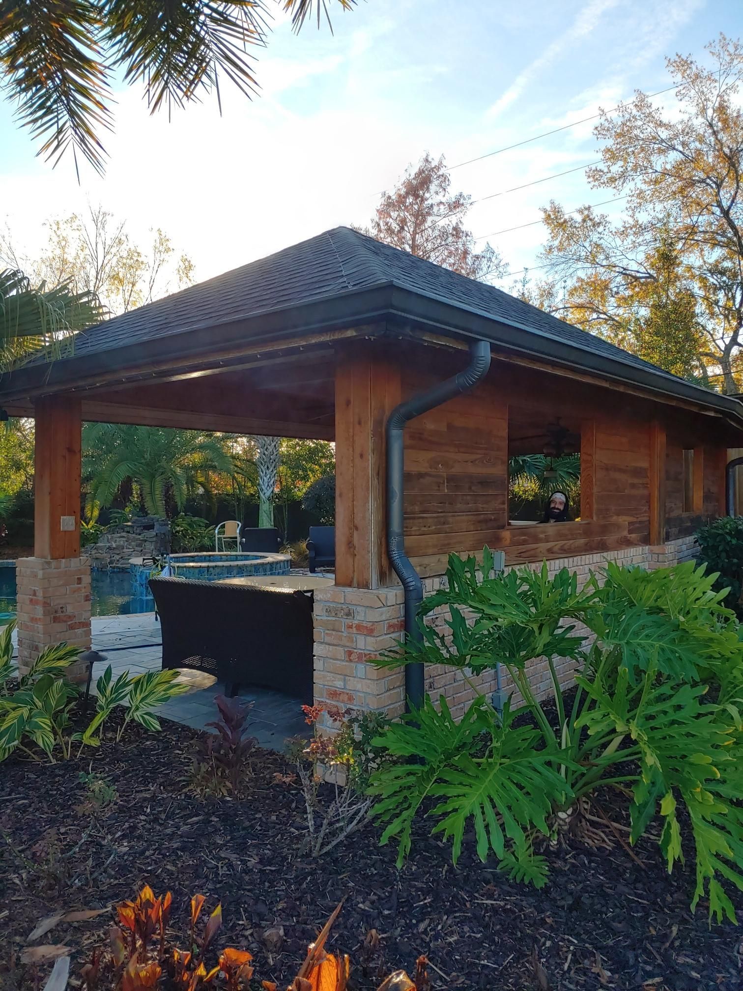 A wooden pavilion with a roof next to a pool and a fire pit.