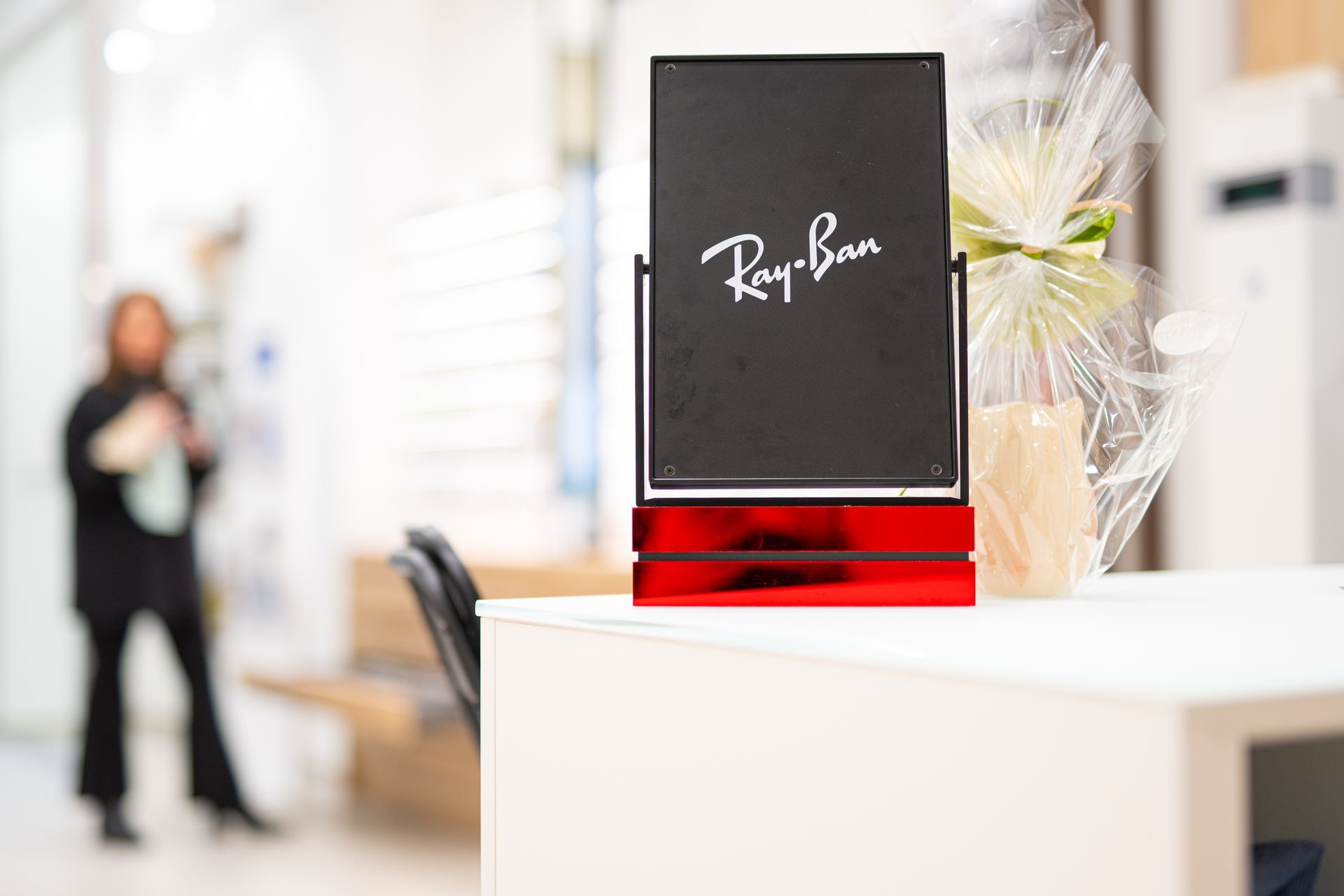 Esposizione di Ray-Ban: insegna nera con logo su base rossa, in un negozio di occhiali. Una persona è sfocata sullo sfondo.