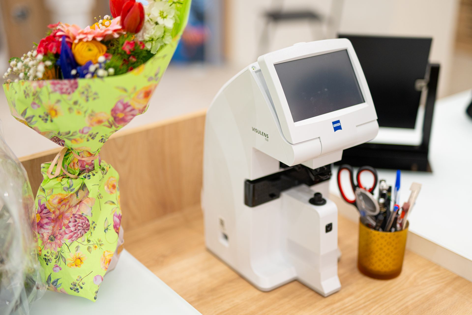 Un apparecchio per l'esame della vista accanto a un mazzo di fiori e un portapenne su una scrivania di legno.