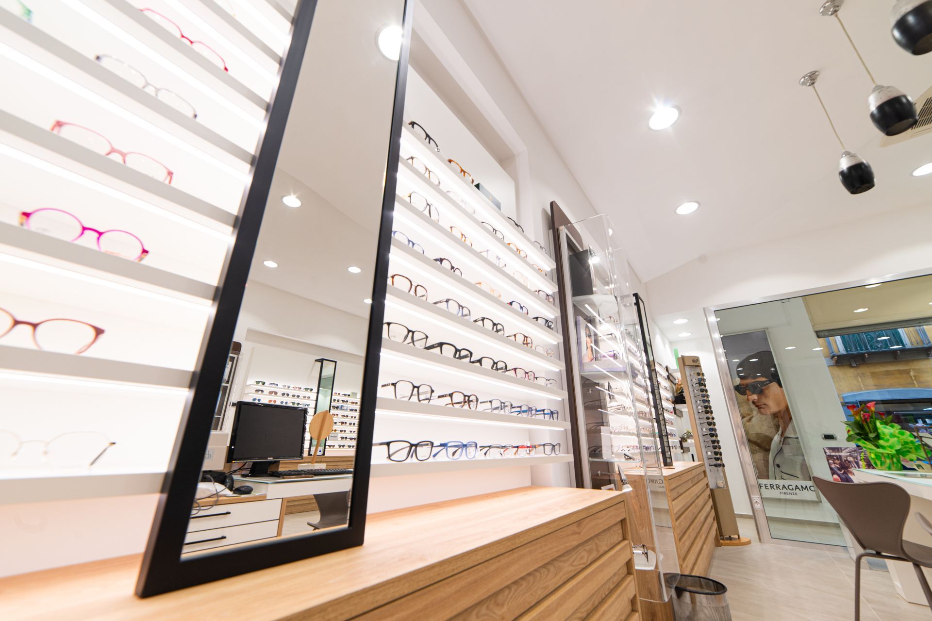Interno di un negozio di ottica con occhiali in esposizione. Un grande specchio riflette l'interno del negozio, con una scrivania e delle sedie visibili.