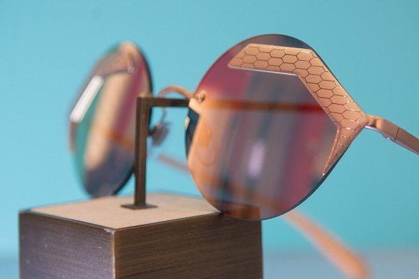 Occhiali da sole color oro rosa con dettagli a nido d'ape, esposti su un supporto, lenti colorate, su sfondo blu.