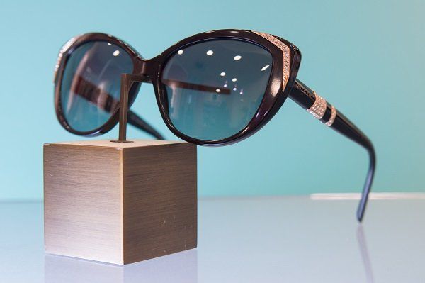 Occhiali da sole neri con dettagli gioiello su un supporto dorato, su uno sfondo blu.