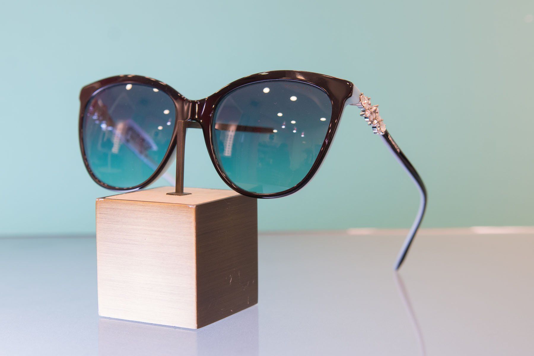 Occhiali da sole neri con lenti blu-verdi su un supporto di legno su uno sfondo azzurro.