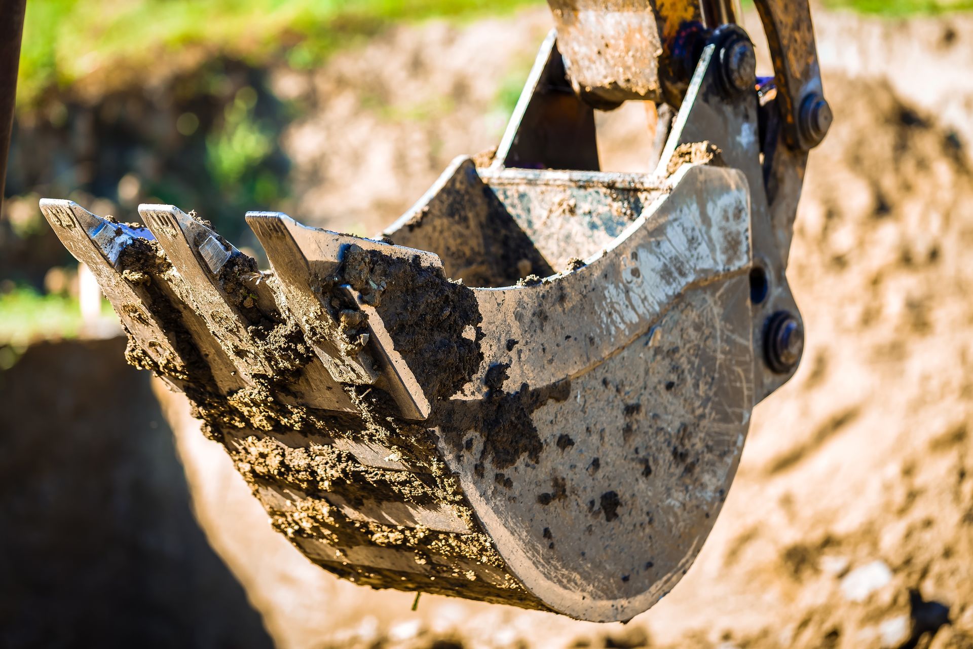 Close-up of a backhoe bucket filled with mud, against a background of a construction site.