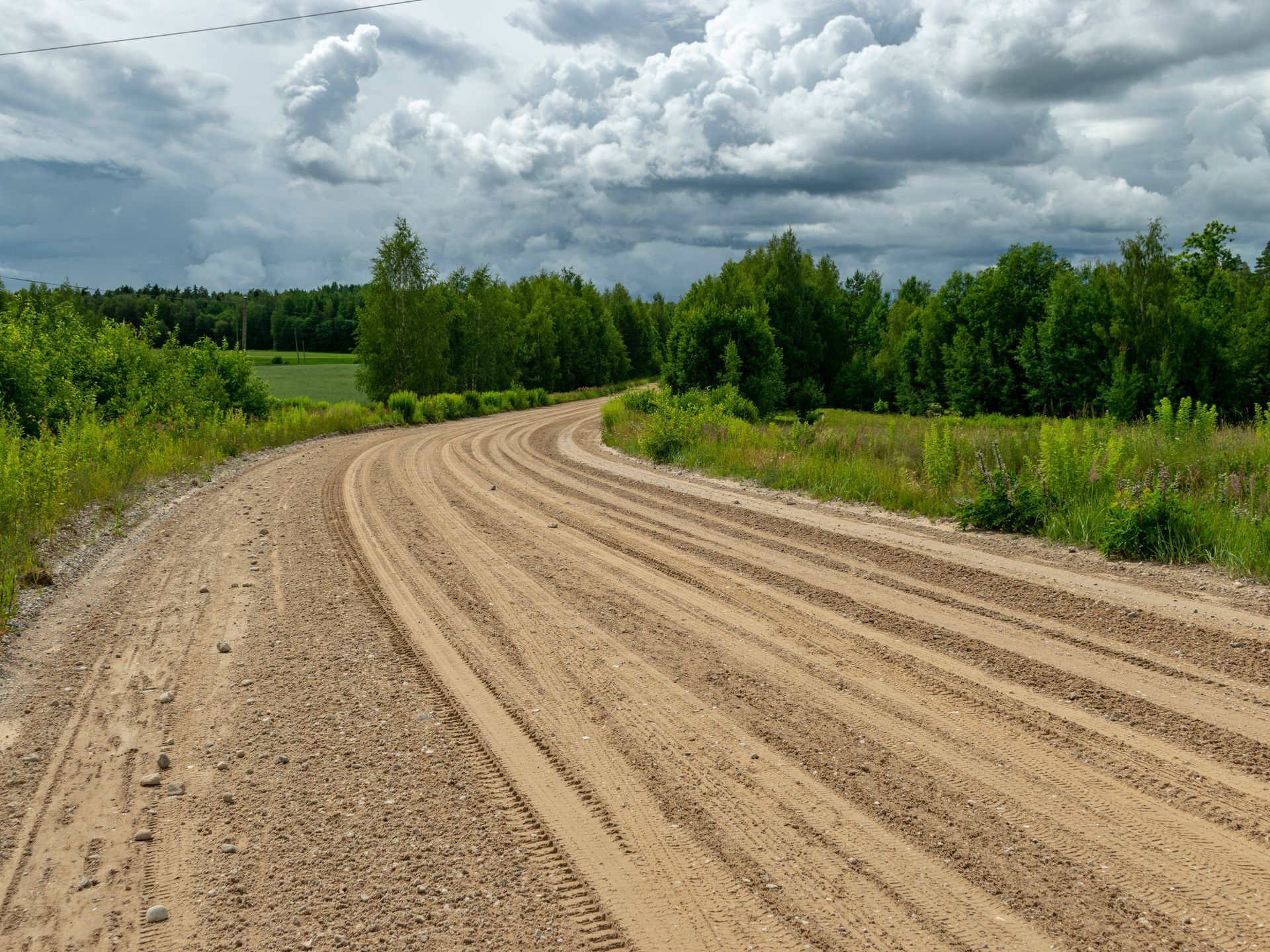 Dirt road curves through a grassy field, lined by green trees under a cloudy sky.