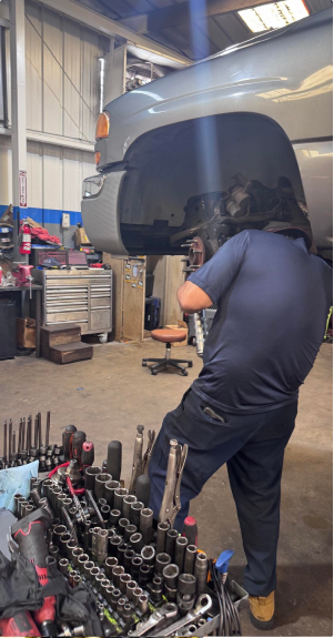 Mechanic in blue uniform at auto shop selects tool from toolbox, working on raised truck. | Allen's Automotive Service
