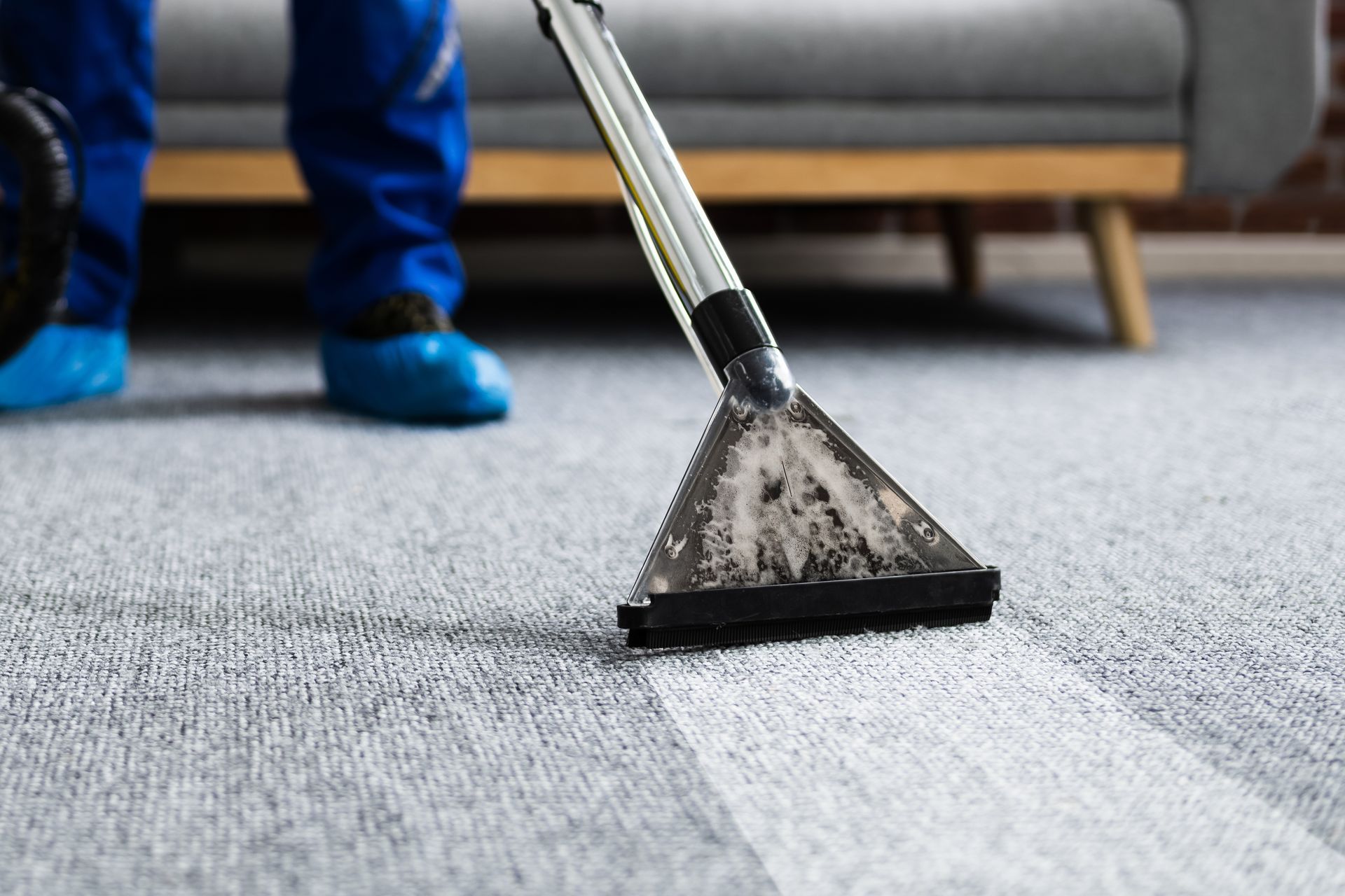 Person in blue coveralls cleaning a light gray carpet with a steam cleaner; a white strip is left in the carpet.