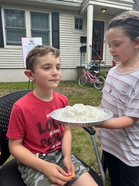 A boy in a red shirt is sitting in a chair while a girl holds a plate of food in front of him.