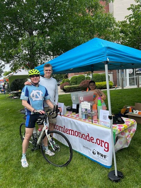 Two men are standing next to a bicycle in front of a lemonade stand.