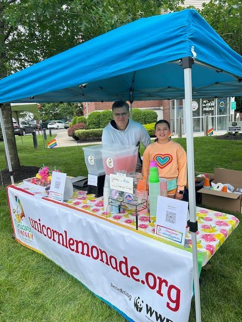 A man and a girl are standing behind a unicorn lemonade stand.