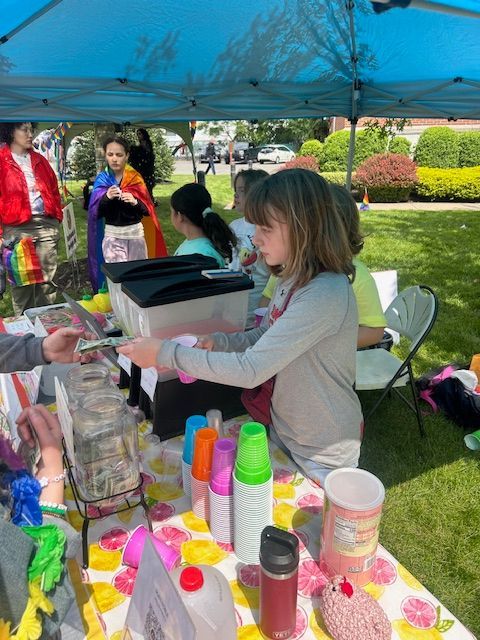 A little girl is standing at a table selling drinks.