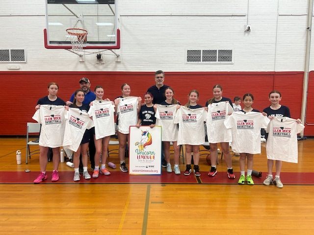 A basketball team stands on a gym court holding up their white team shirts with a sign in the center.