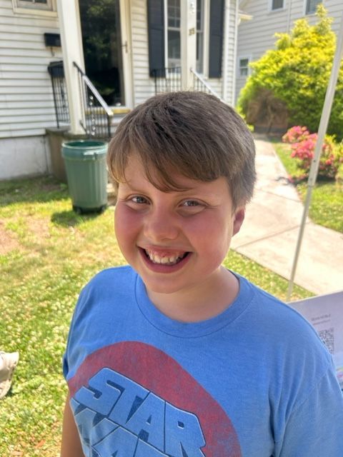 A young boy wearing a blue star wars t-shirt is smiling in front of a house.