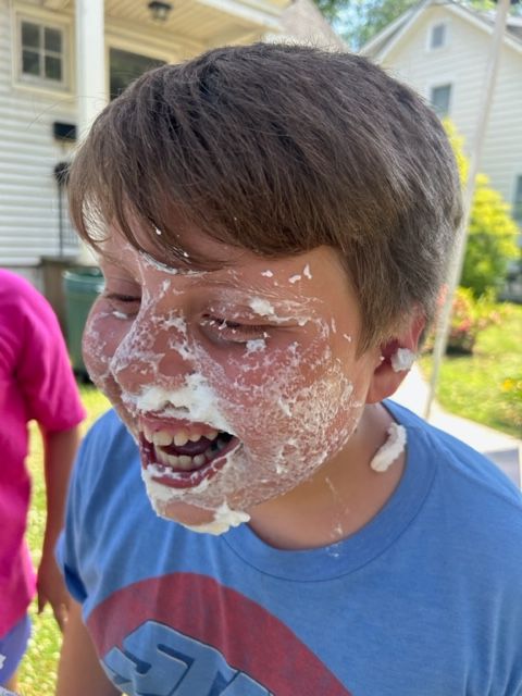 A boy with whipped cream on his face is wearing a blue shirt with the letter s on it