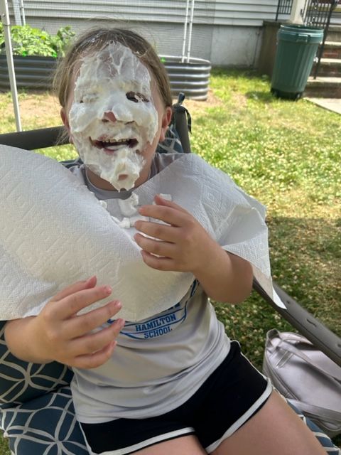 A little girl with whipped cream on her face is sitting in a chair holding a paper towel.