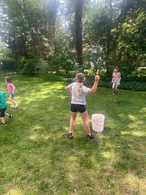 A girl is throwing a ball into a bucket in a yard.