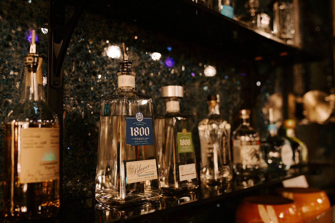 A row of bottles of alcohol are lined up on a bar shelf.