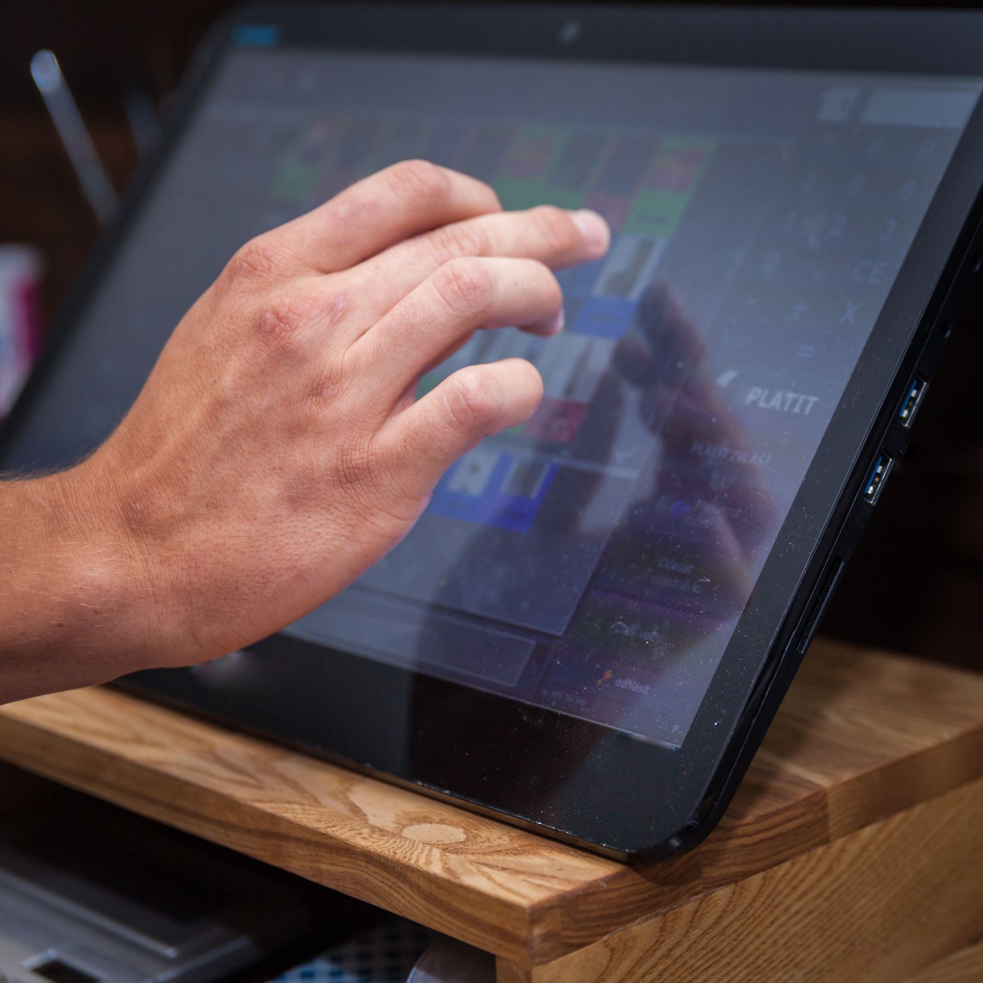 Hand interacting with a touch screen, likely a point-of-sale system, on a wooden stand.