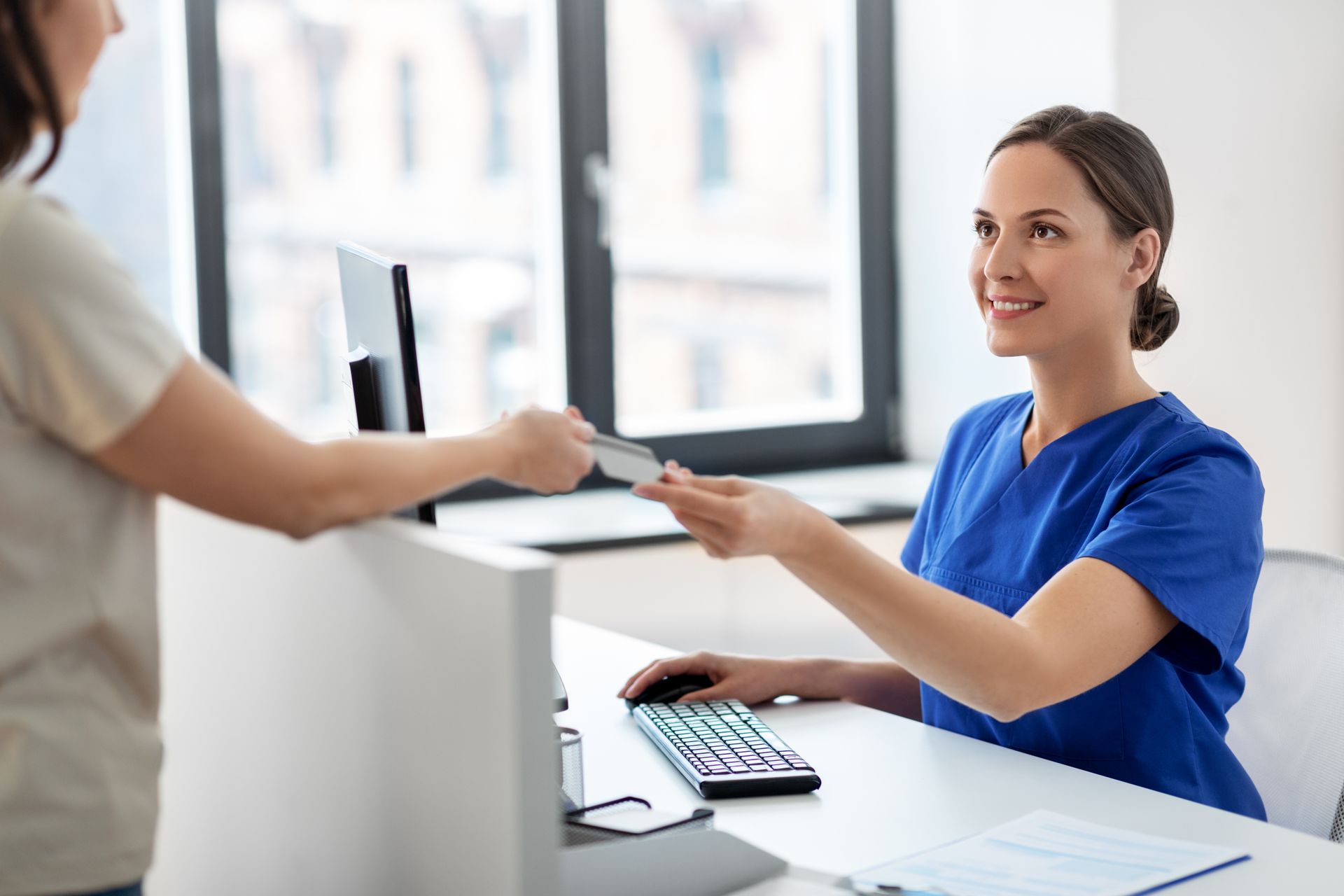 Woman in blue scrubs smiles, receiving card from patient at a reception desk.