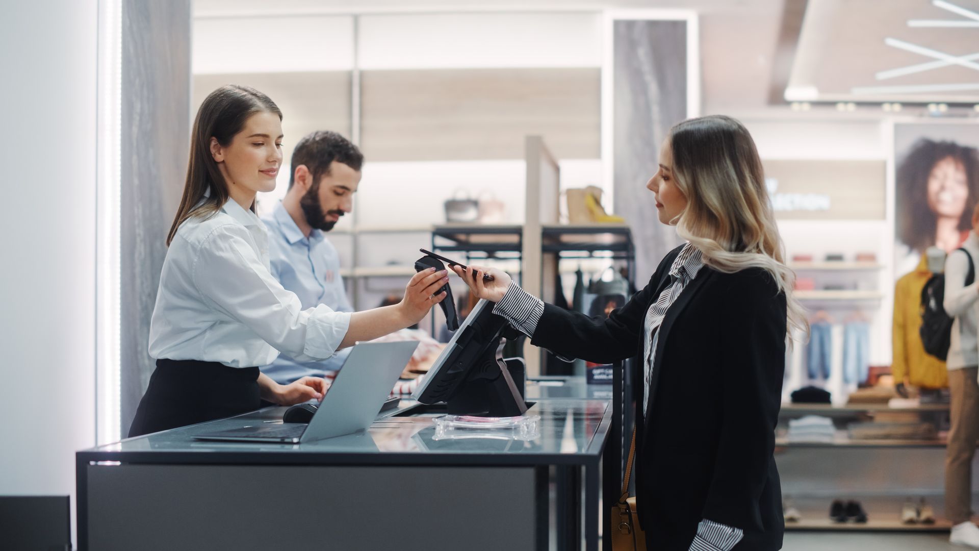 Woman paying at a store counter. A cashier hands over a shopping bag. Another person works at the register.