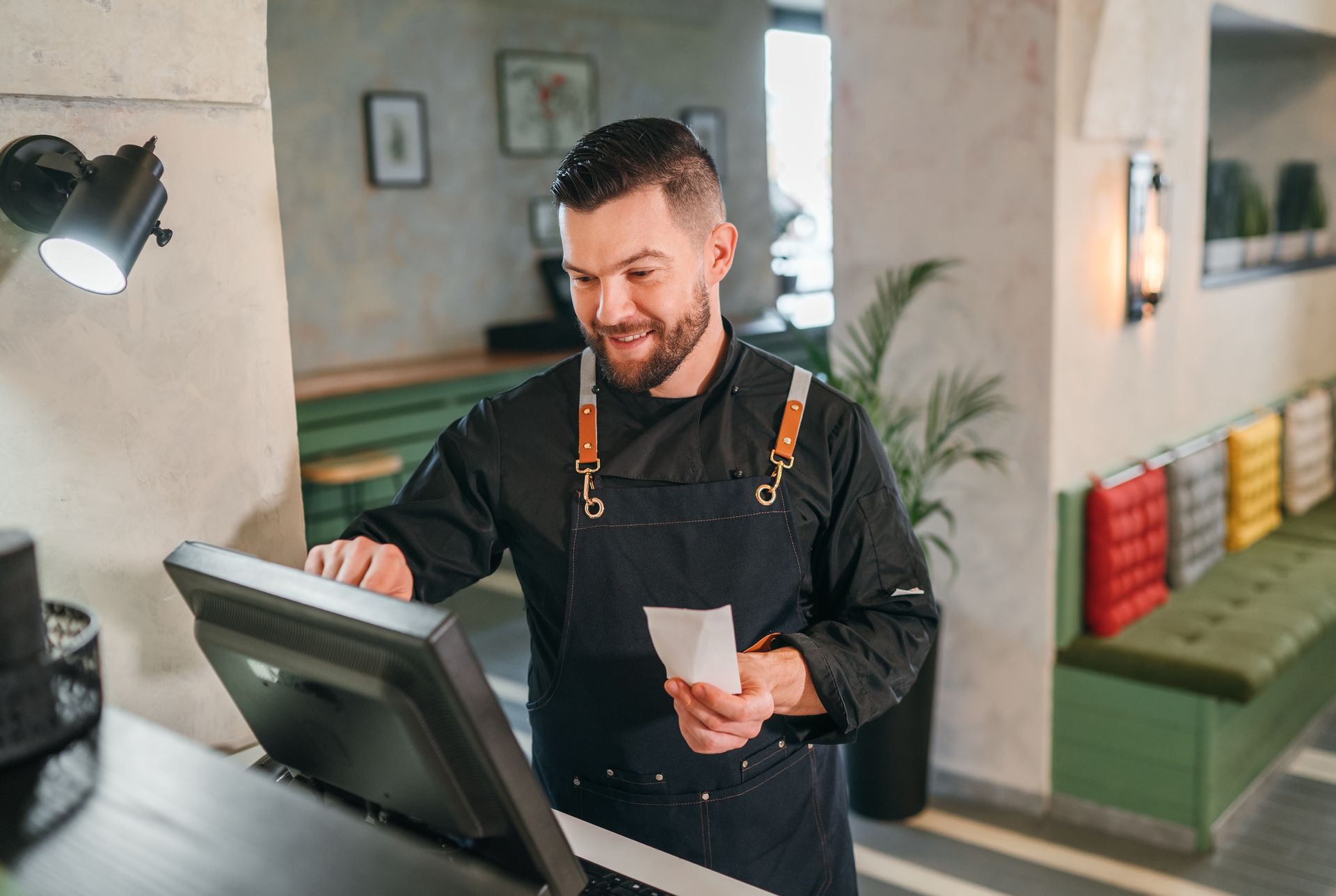 Man in black apron at a cash register, smiling.