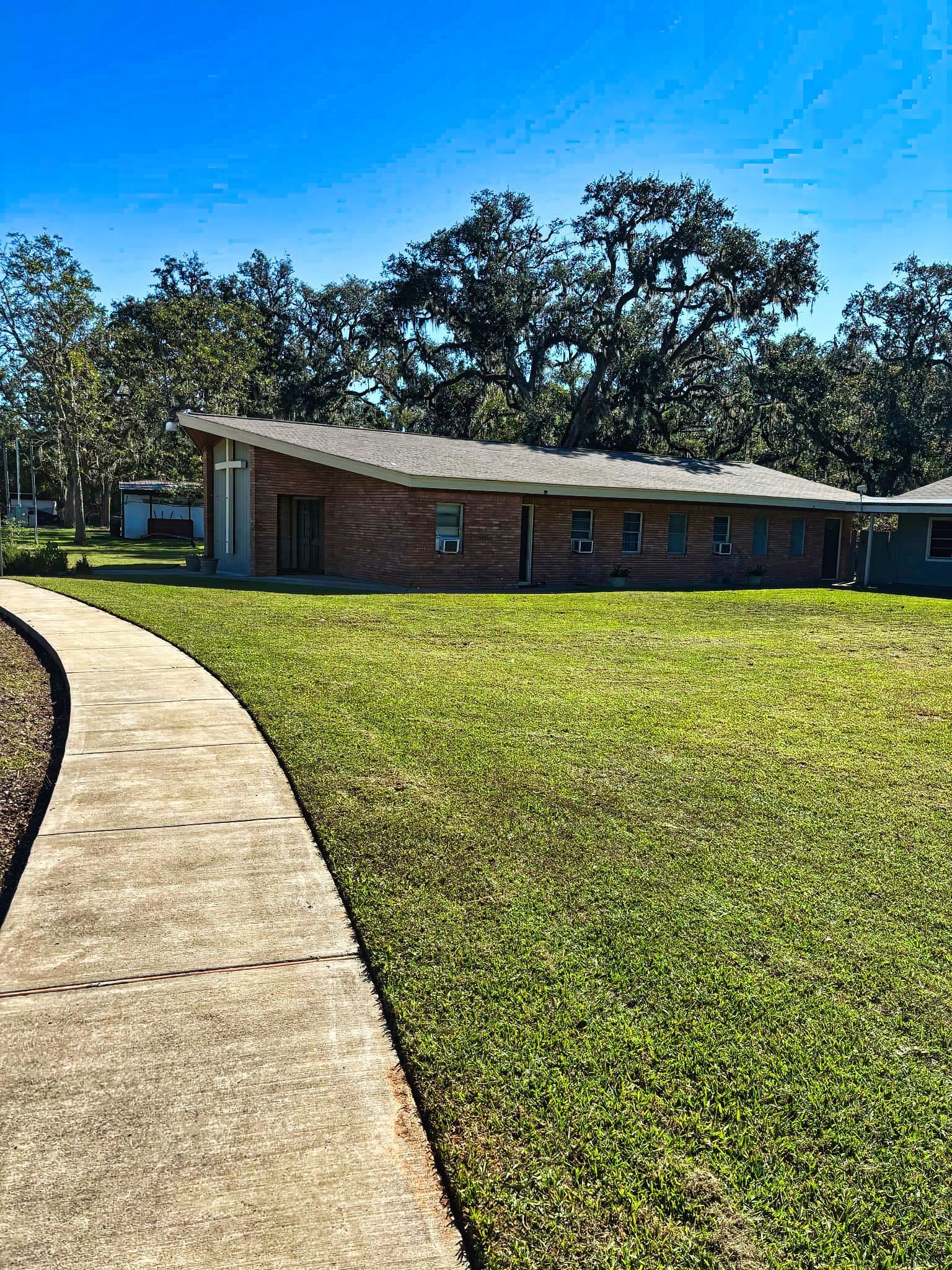A low brick building with a curved roof sits on a grassy lawn, with a sidewalk leading toward it. Trees line the background.