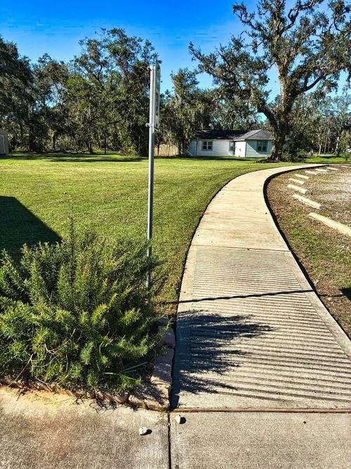 A curved sidewalk leads to a small house, passing a green bush and a metal pole. The path casts circular shadows.
