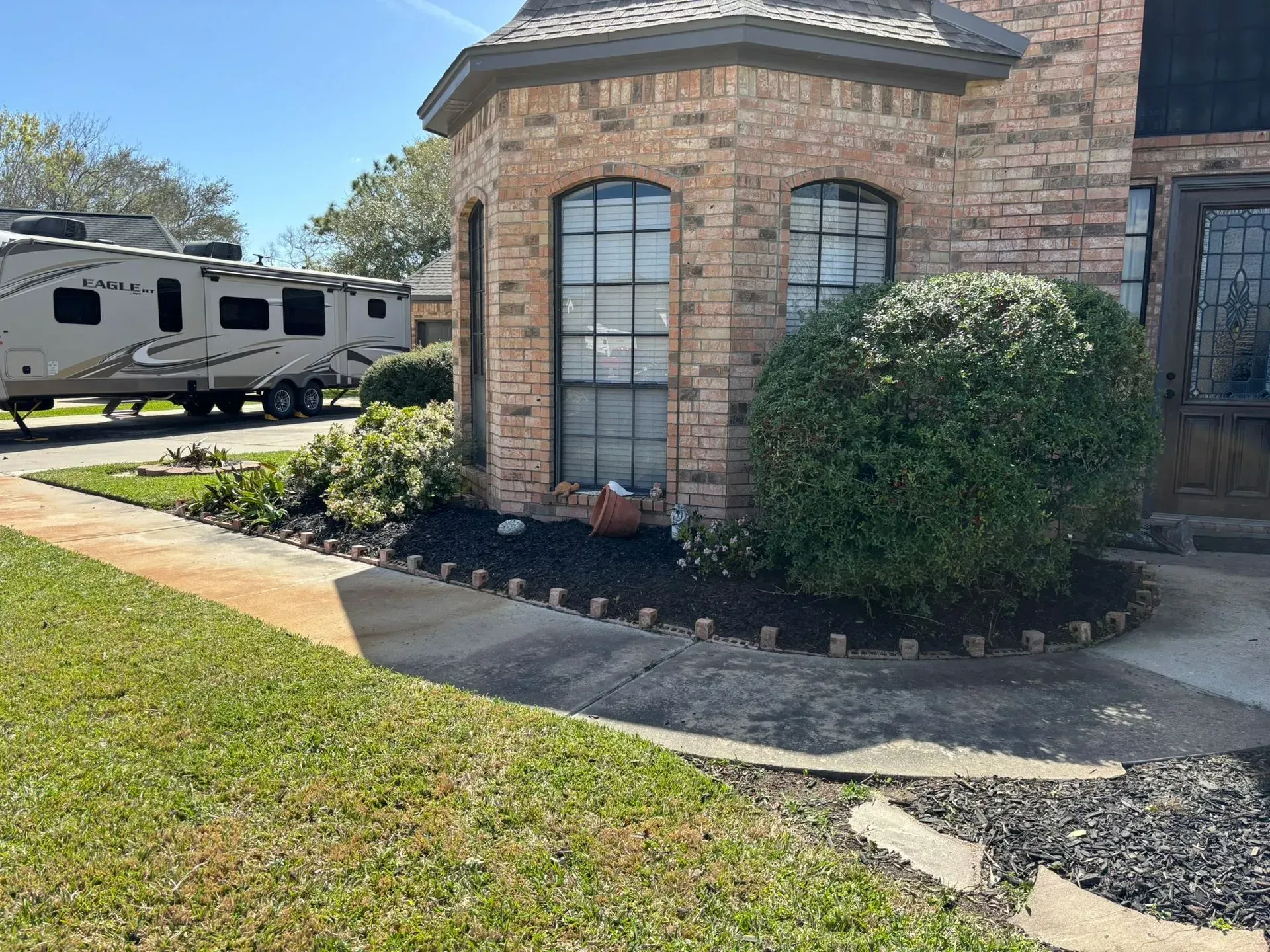 A brick house with an RV parked in the driveway. Green grass in front and a garden bed with mulch and shrubs.