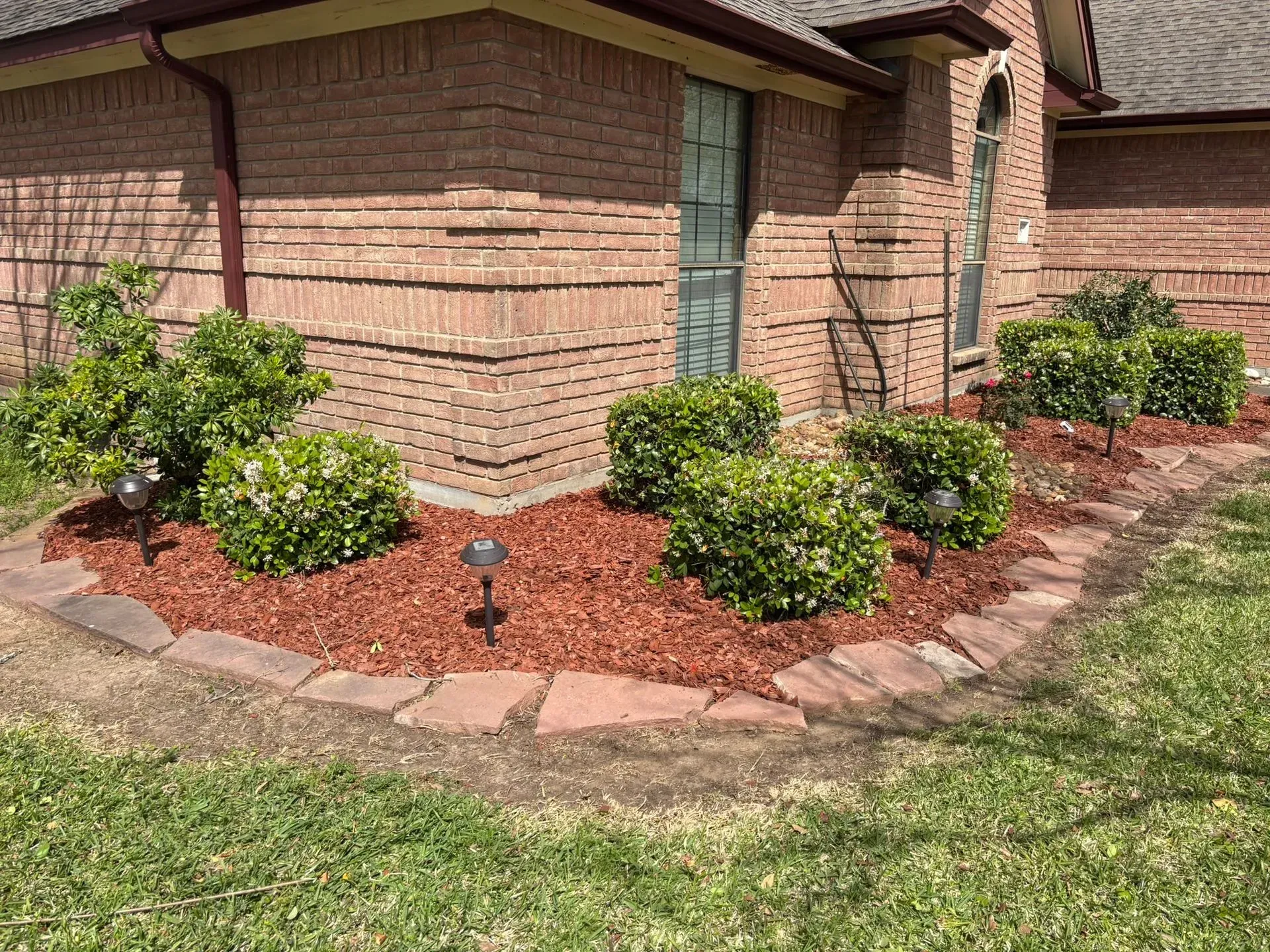 The bed is lined with red mulch and stone, and contains green bushes and solar lights.
