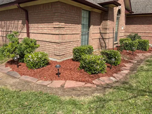 Brick house with flower bed edged with stones, filled with red mulch, and several green bushes.