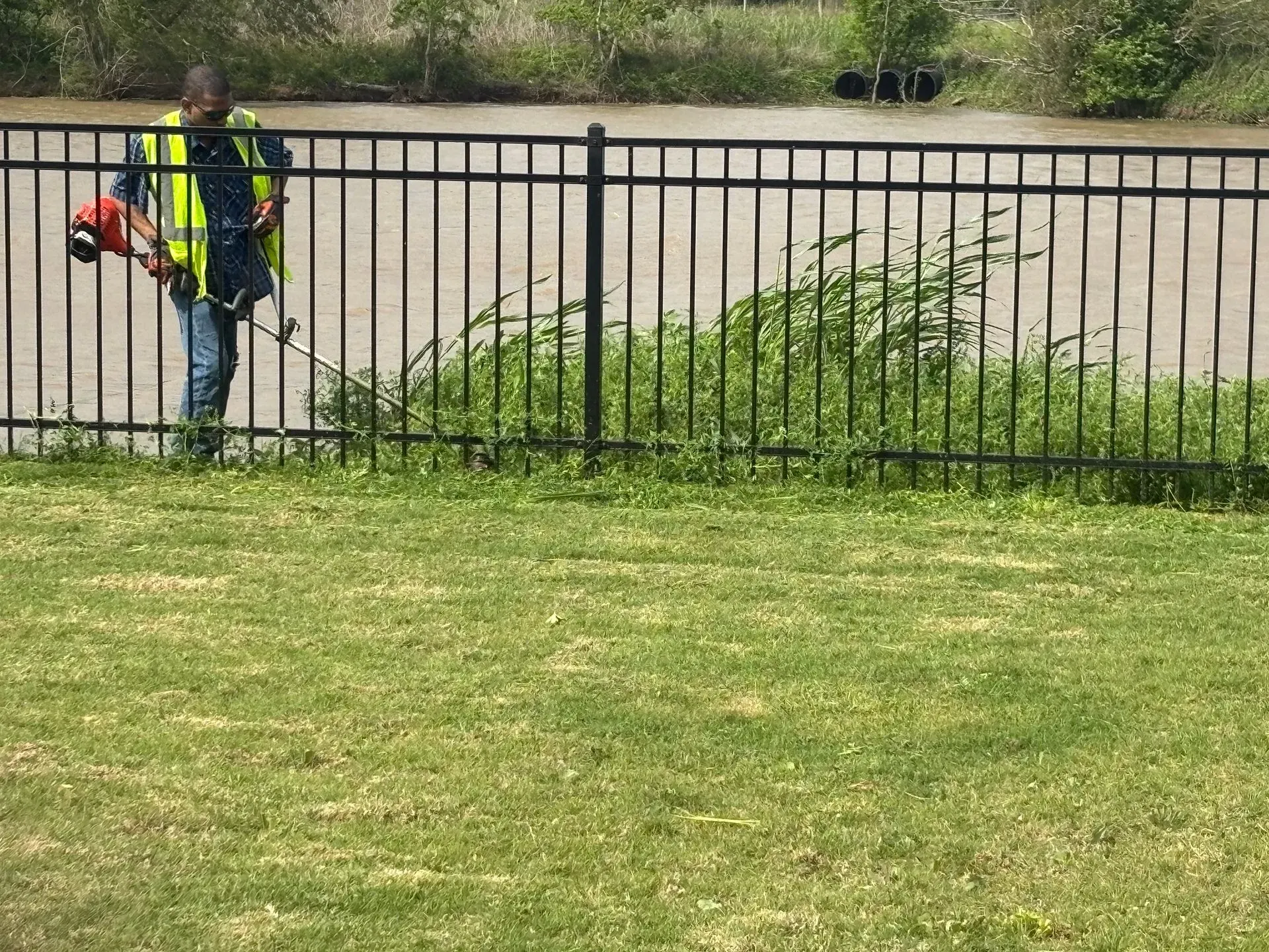 A person in a reflective vest uses a weed wacker along a fence next to water.