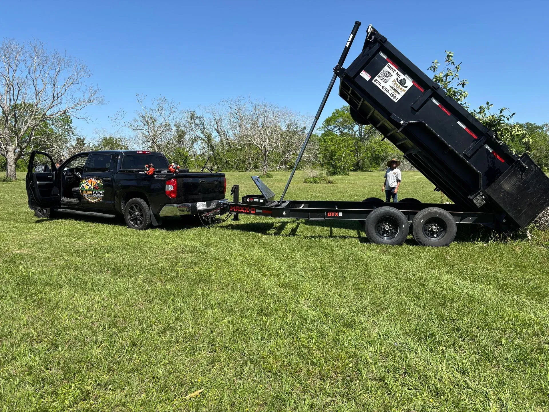 A black pickup truck towing a raised black dump trailer on a grassy field under a blue sky. A person stands near the trailer.