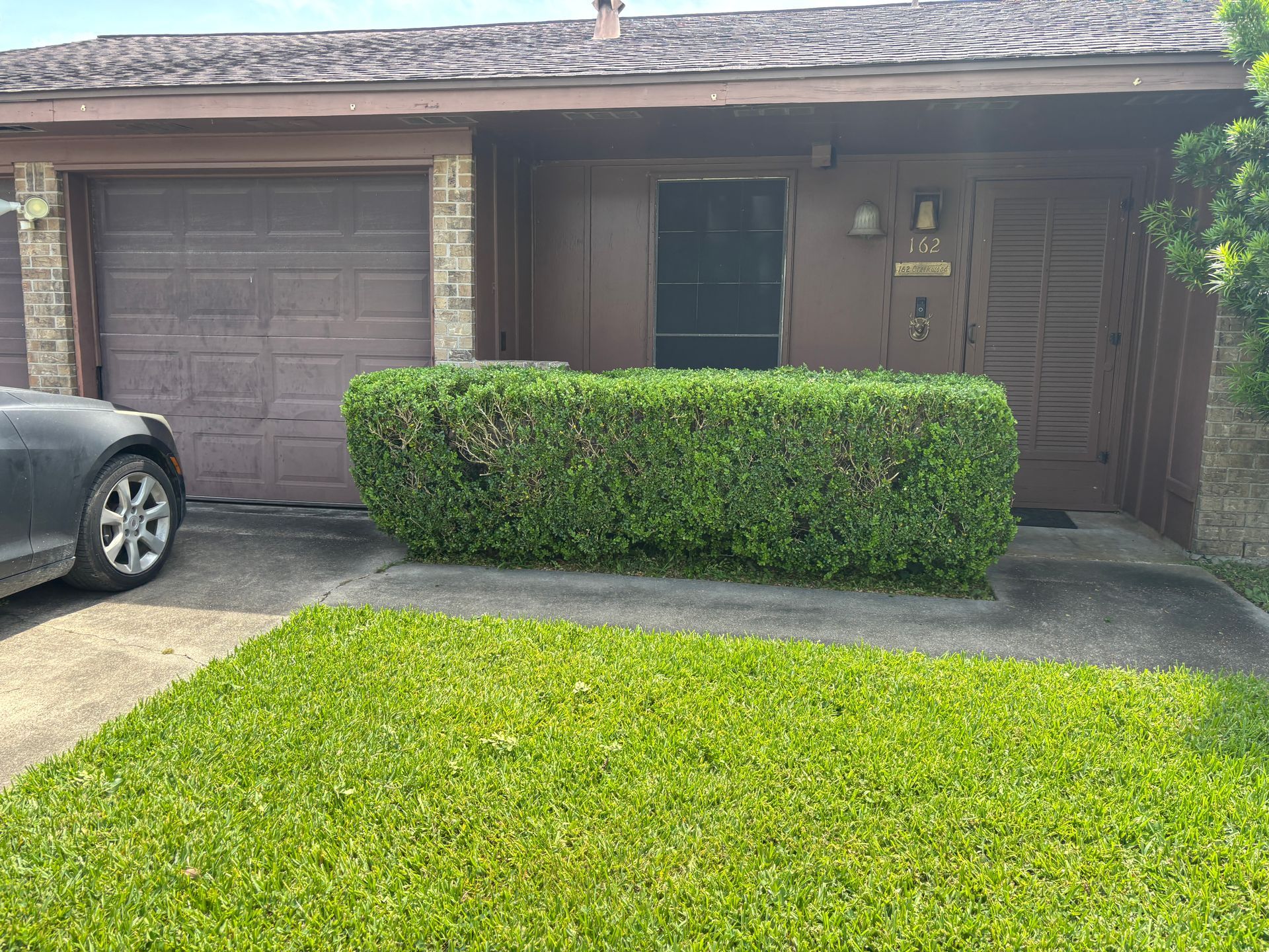 A brown house with a garage and a well-trimmed green hedge in front. A car is parked on the left.