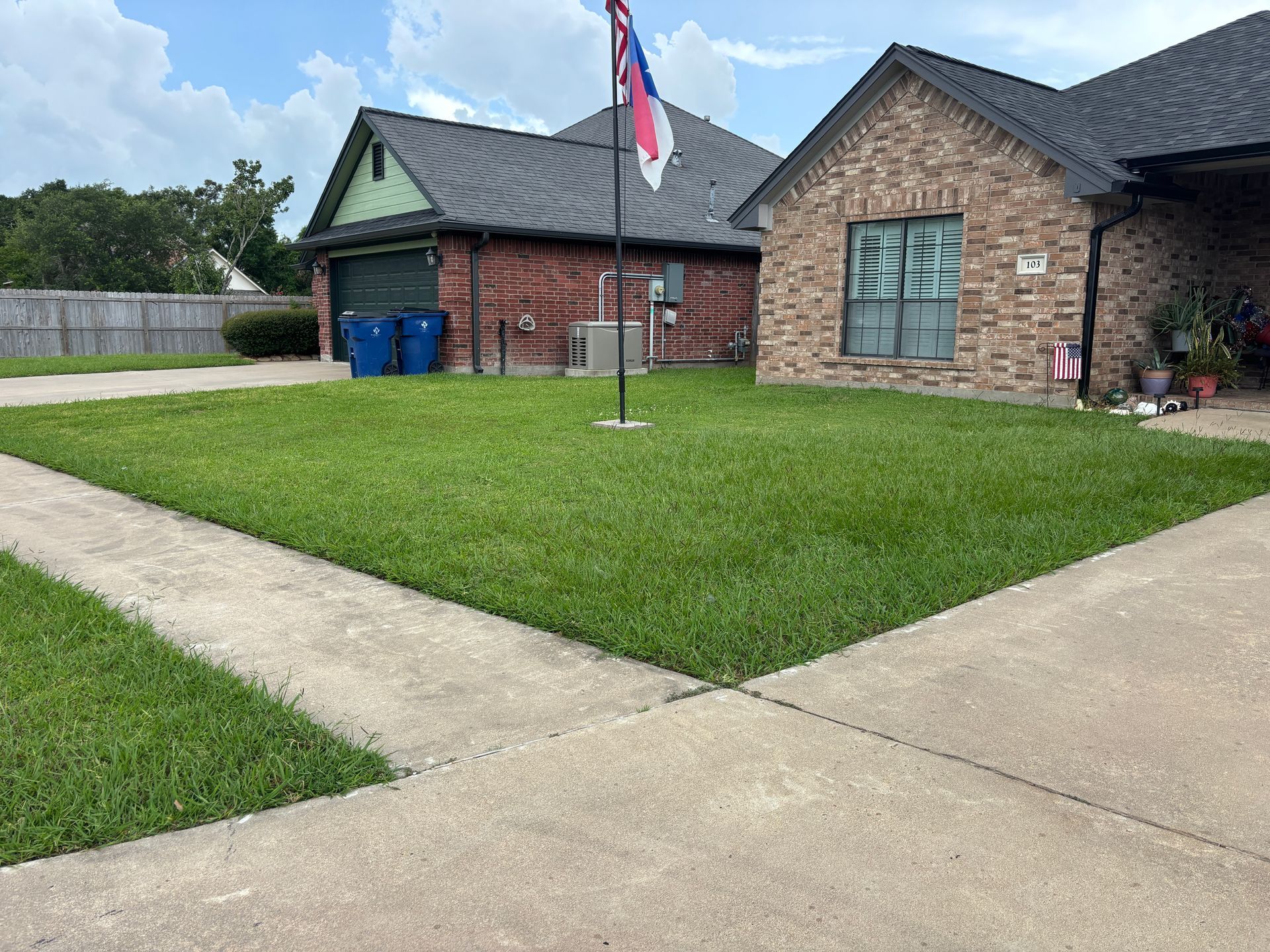 A well-manicured green lawn in front of a brick house with a garage, a sidewalk, and a flag in the yard.
