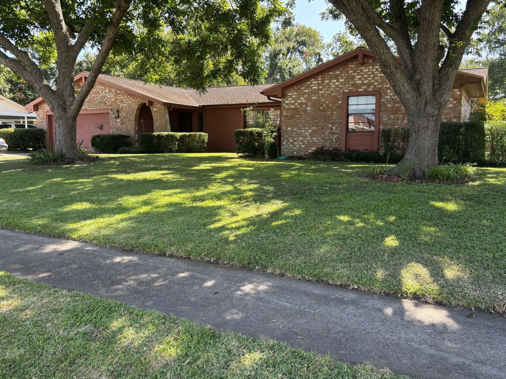 A single-story brick home with a well-manicured lawn, two large trees, and a sidewalk in front.