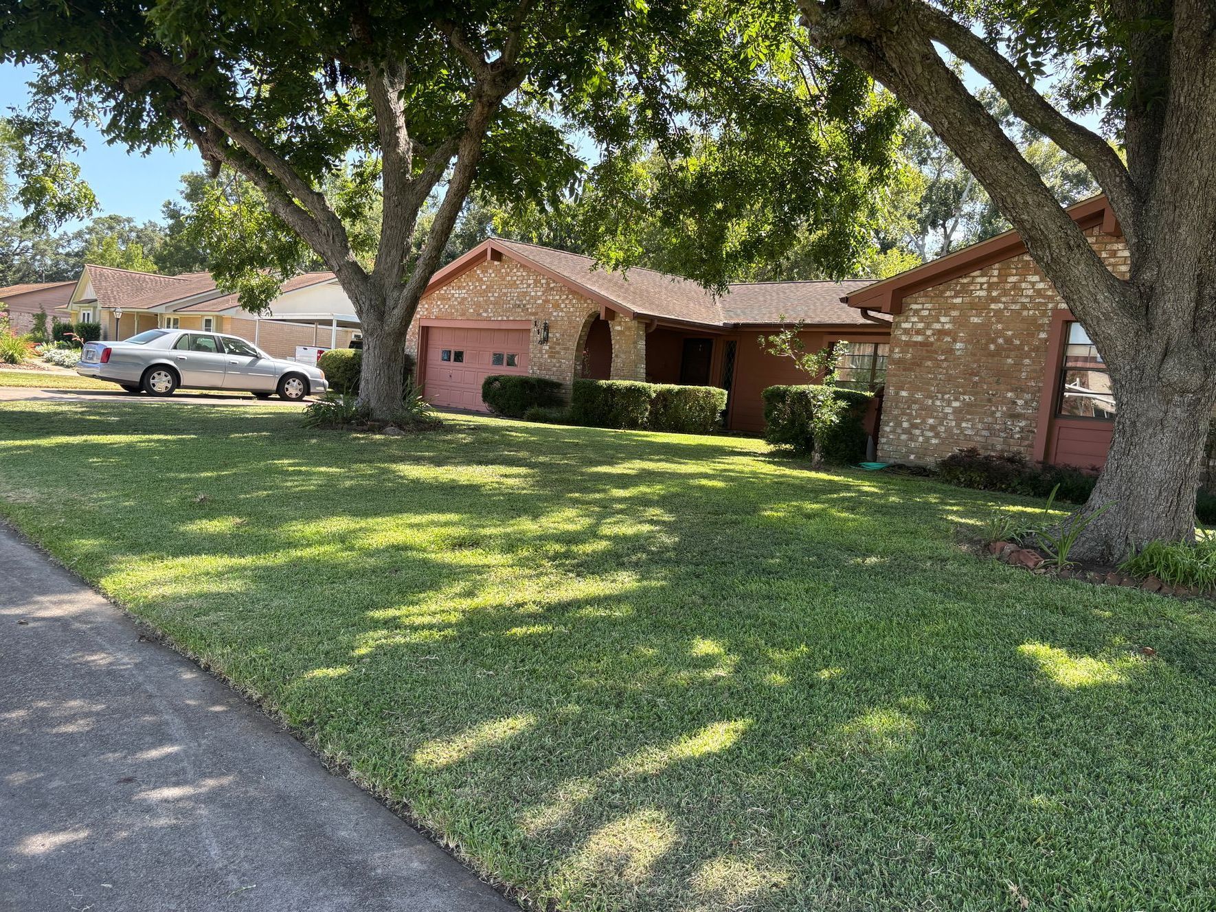 A one-story brick house with a green lawn shaded by trees. A car is parked in front of a neighbor's house.