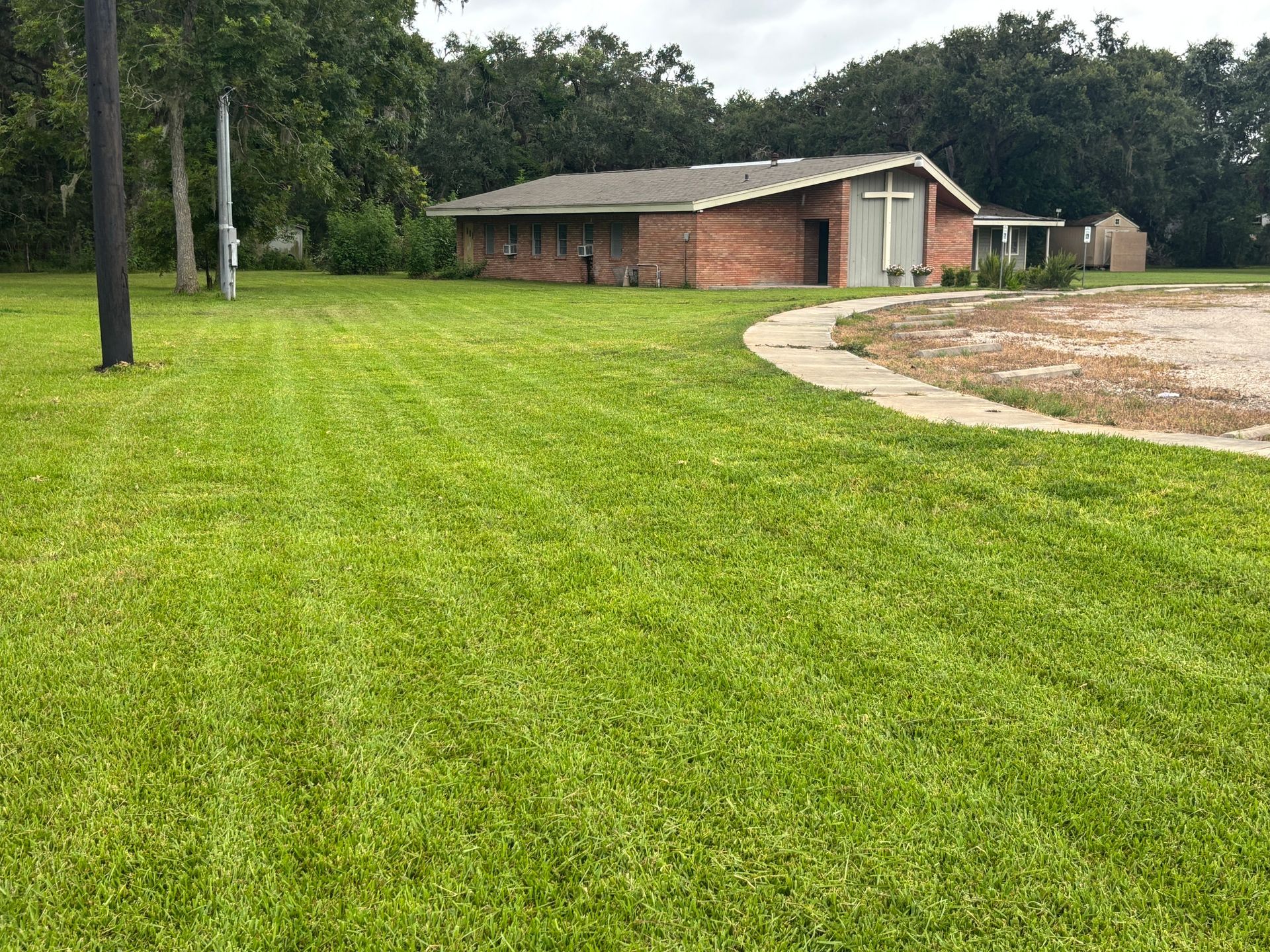 A single-story brick church with a cross above the entrance on a green lawn. A paved path curves toward the church.