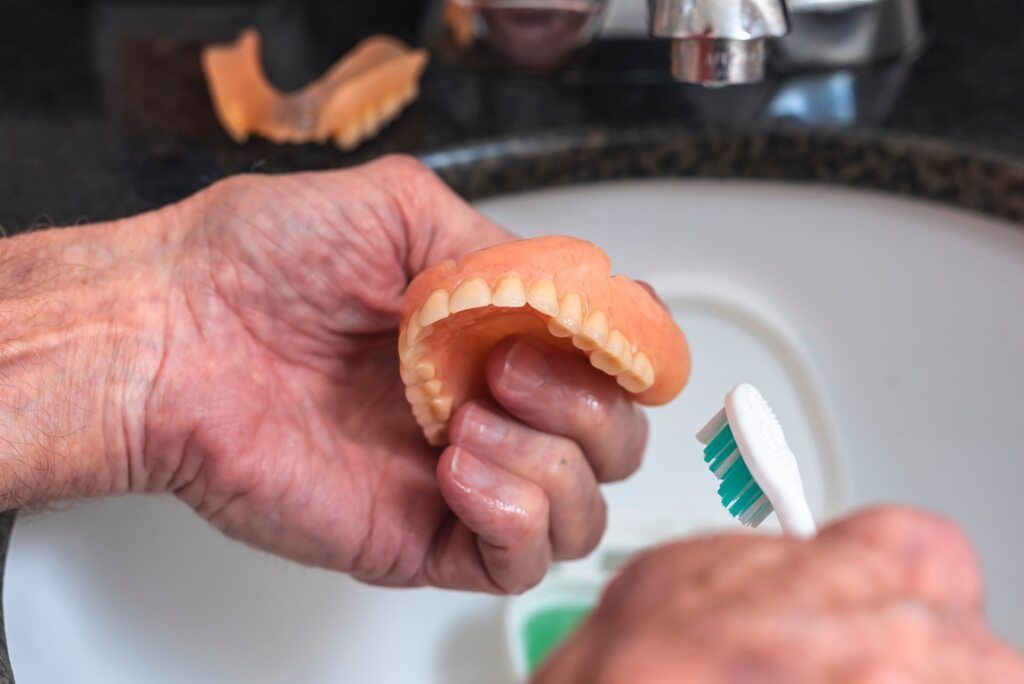 Person Cleans Dentures With a Toothbrush in a Sink — The Denture Care Group Sunshine Coast in Caloundra, QLD