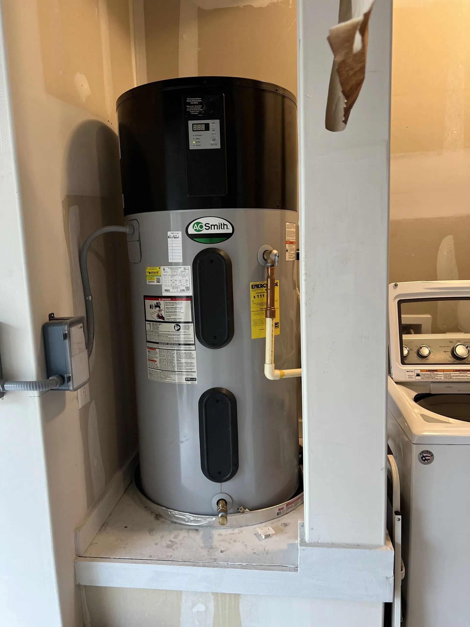 A tall electric water heater in a laundry room, with black top, gray tank, and beige pipes.
