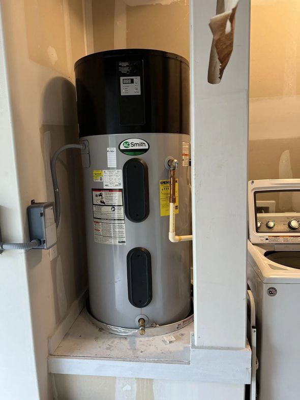 A tall electric water heater in a laundry room, with black top, gray tank, and beige pipes.