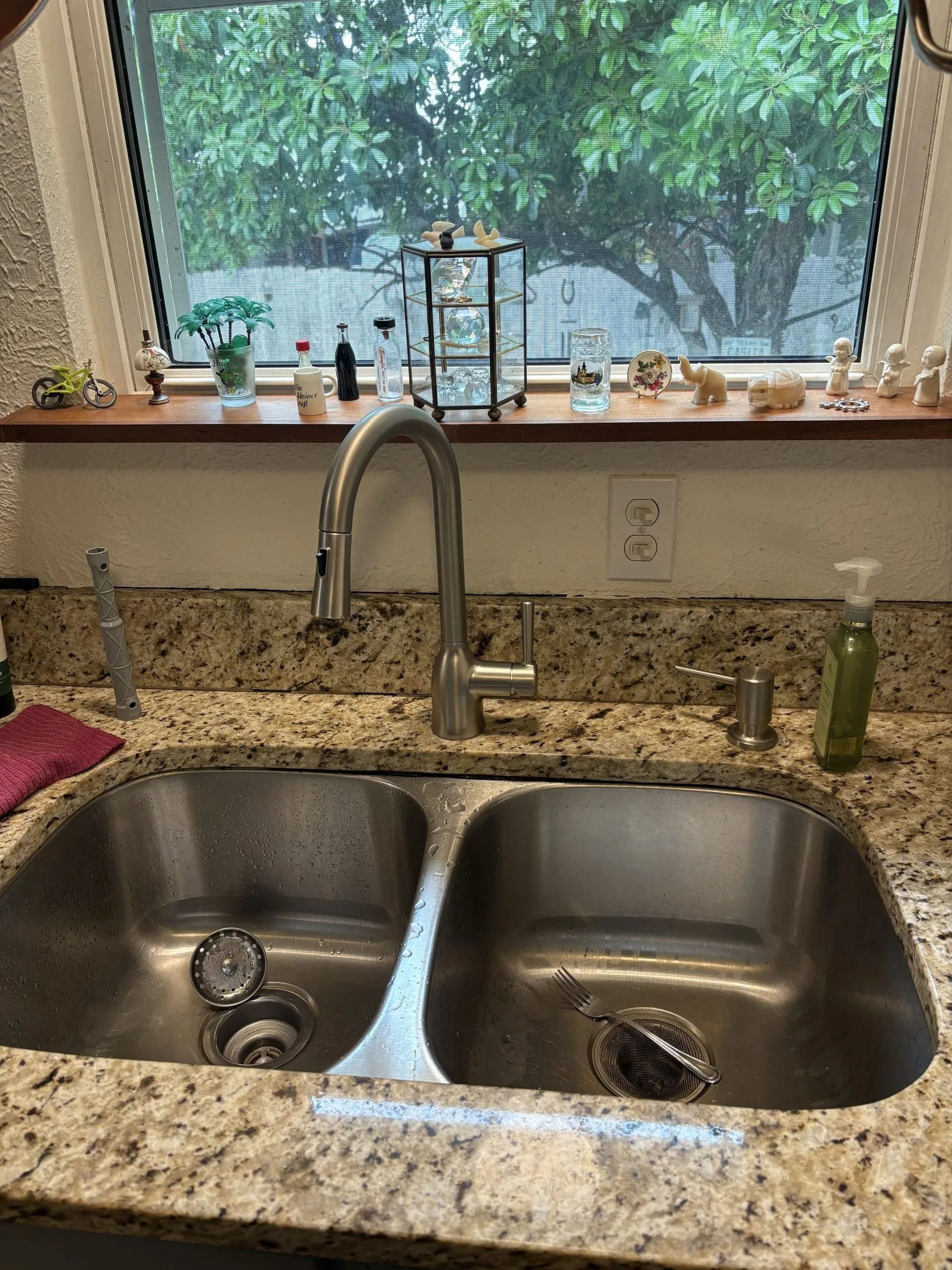 Stainless steel kitchen sink with faucet, granite countertop, window with outdoor view.