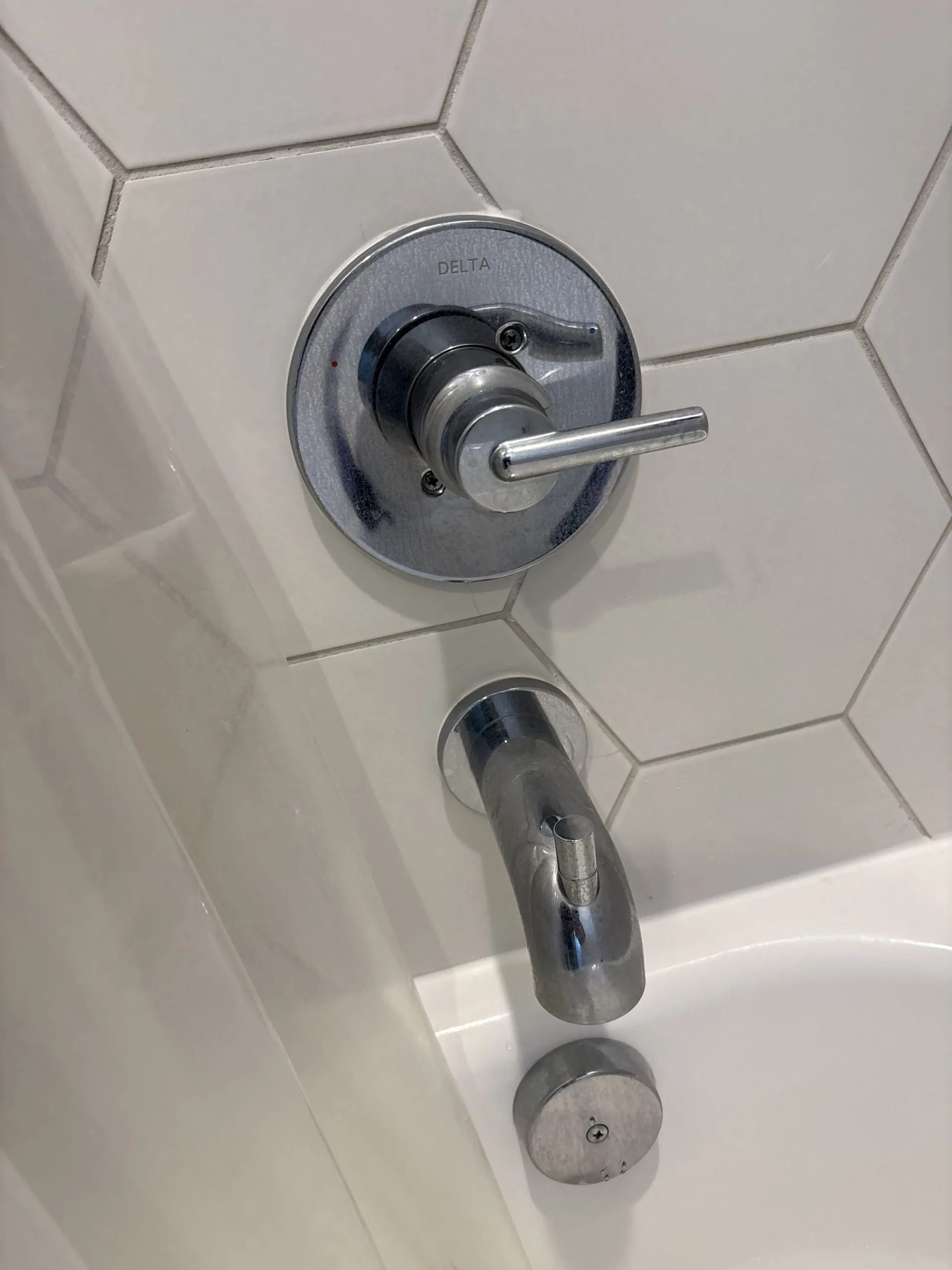 Chrome shower faucet with lever handle, mounted on white hexagon tile above the tub.