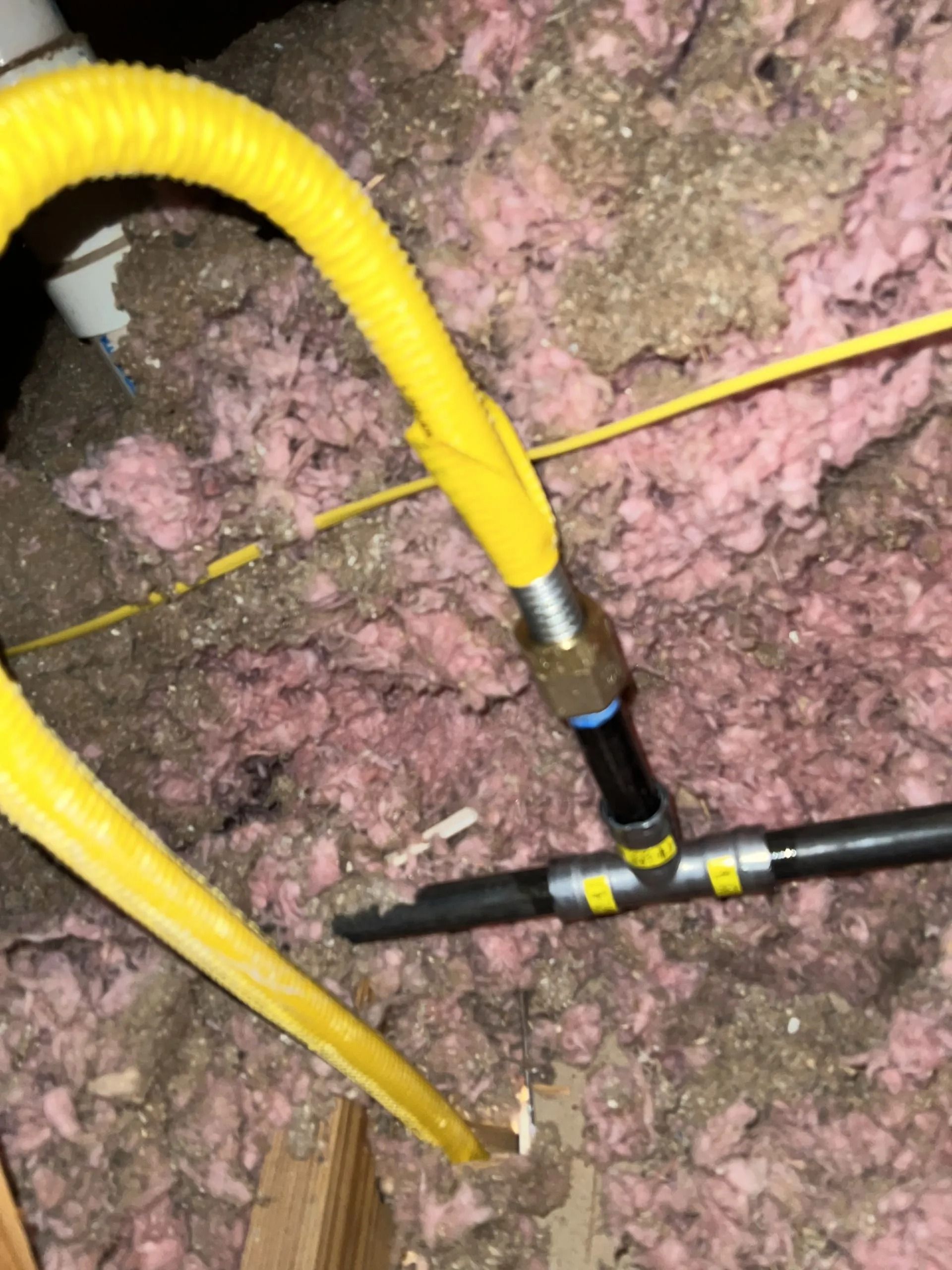 Yellow corrugated gas line connected to black pipes within a dusty insulation-filled space.