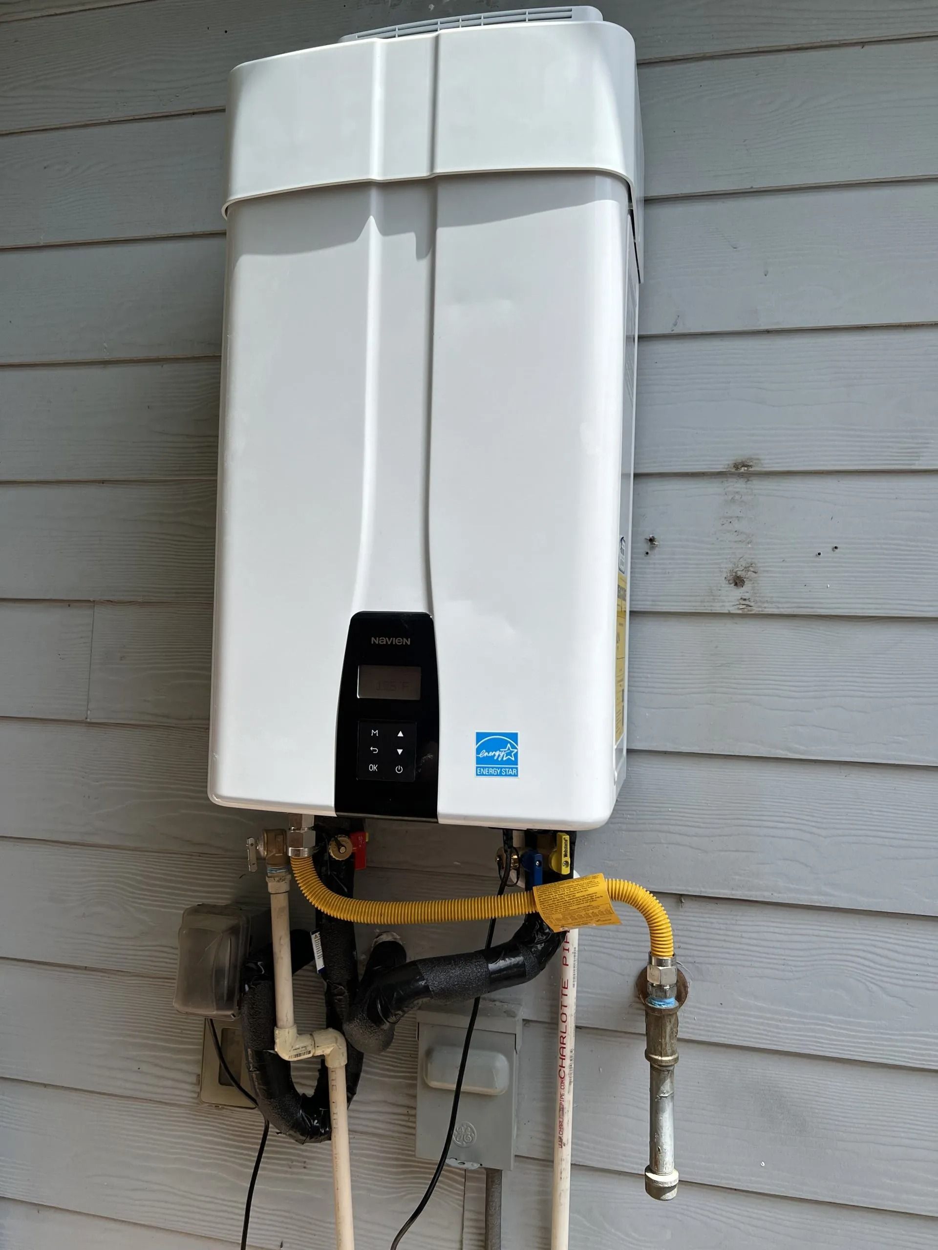 White tankless water heater on a gray wooden wall, connected to gas and water pipes.