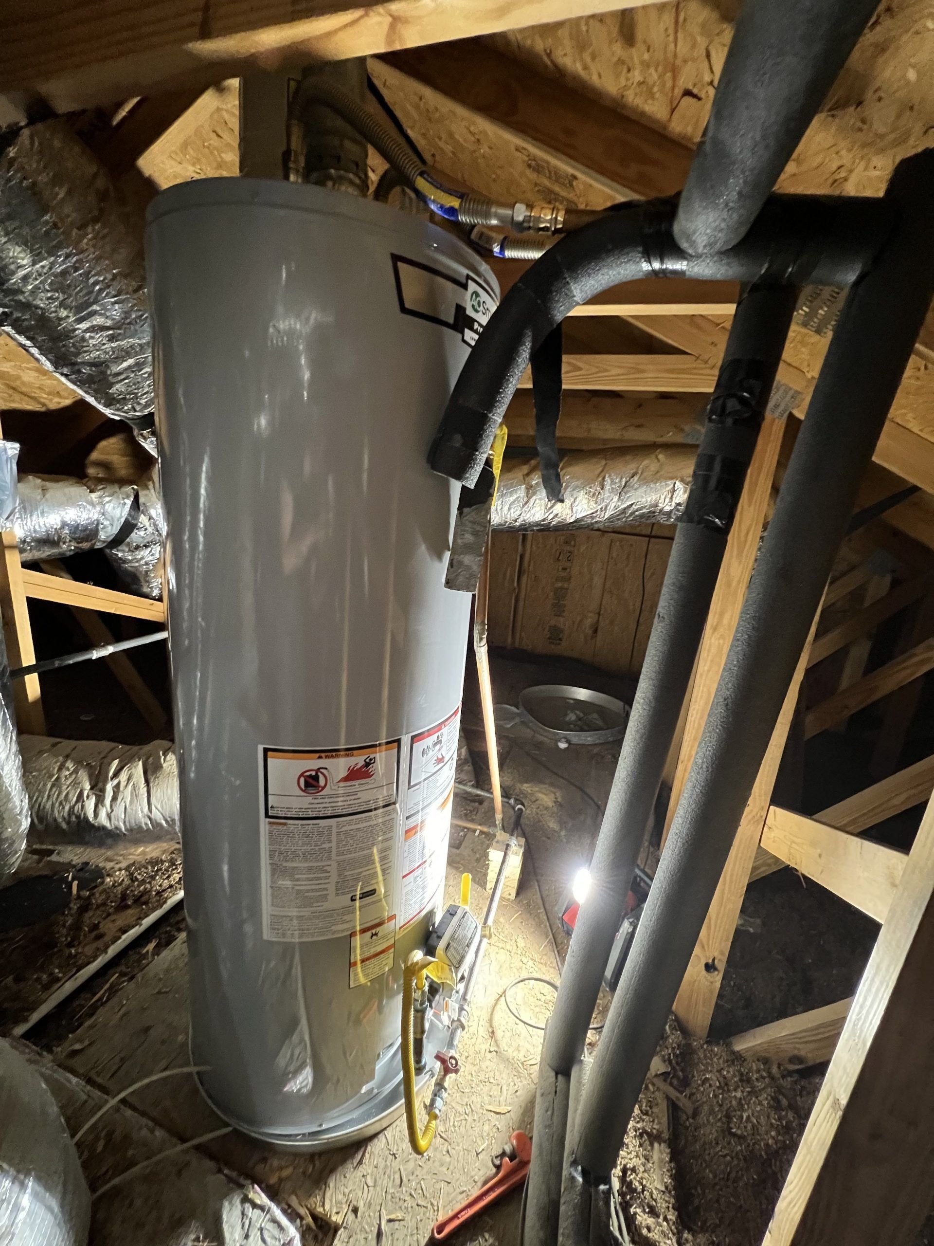 A water heater in an attic with pipes and exposed beams.