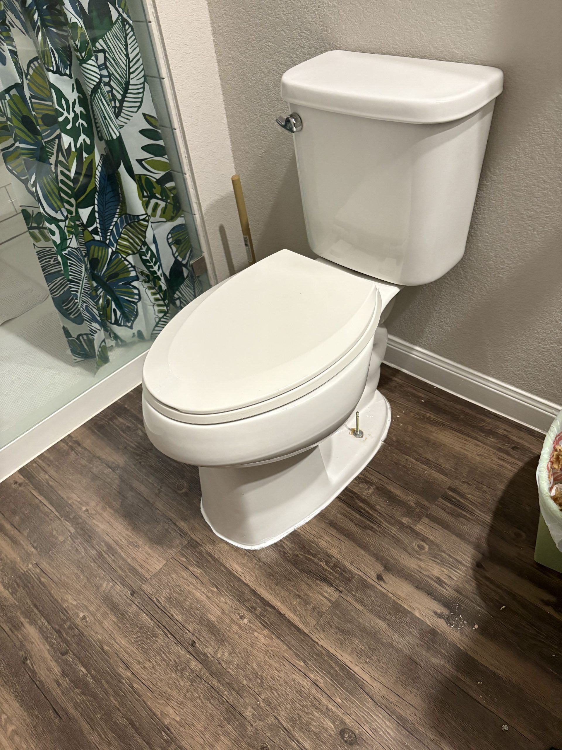 White toilet in bathroom with wood-look flooring. Shower with leaf-patterned curtain.