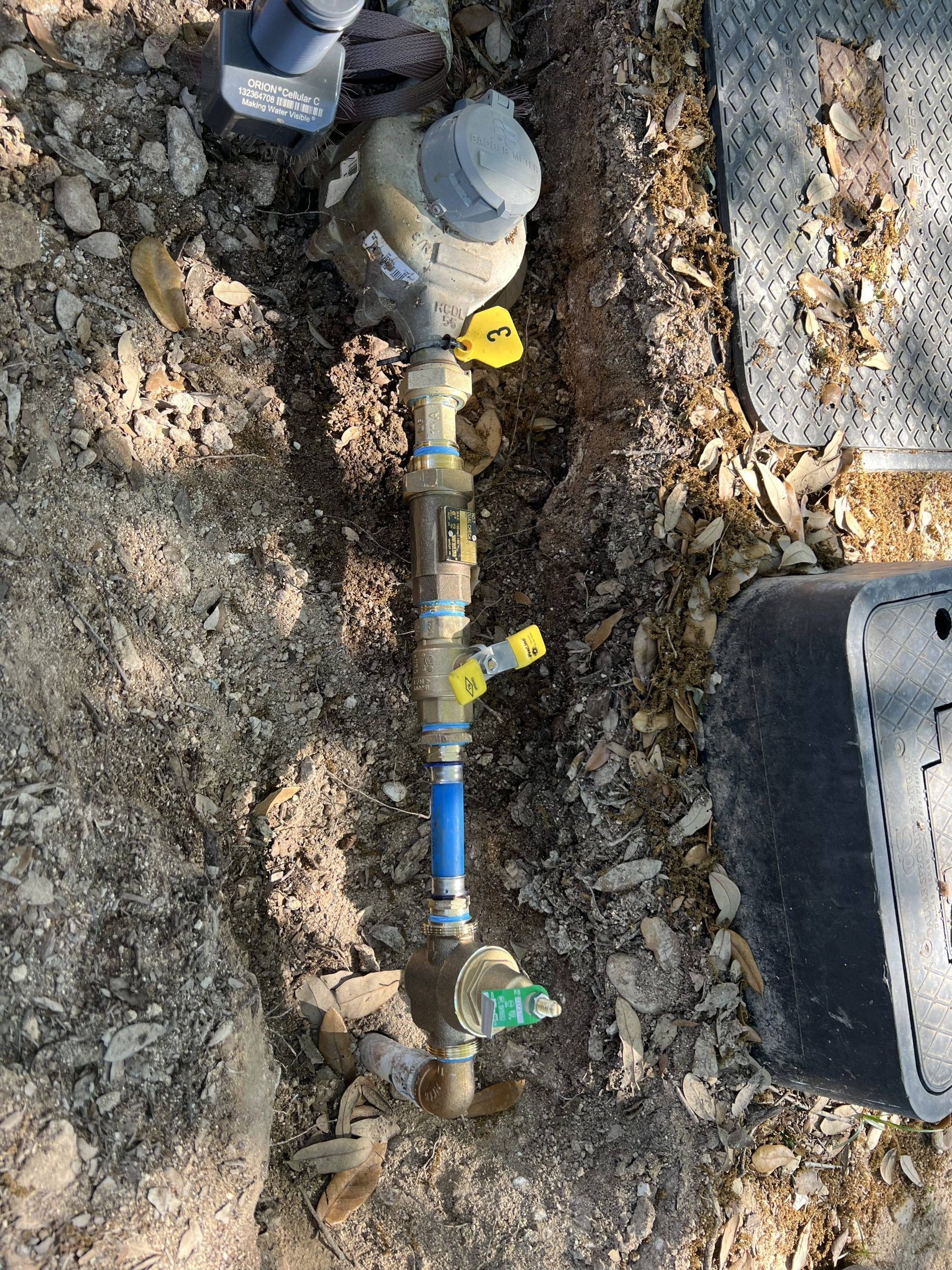 Water meter with pipes, valves, and a blue section, outdoors in dirt and debris.