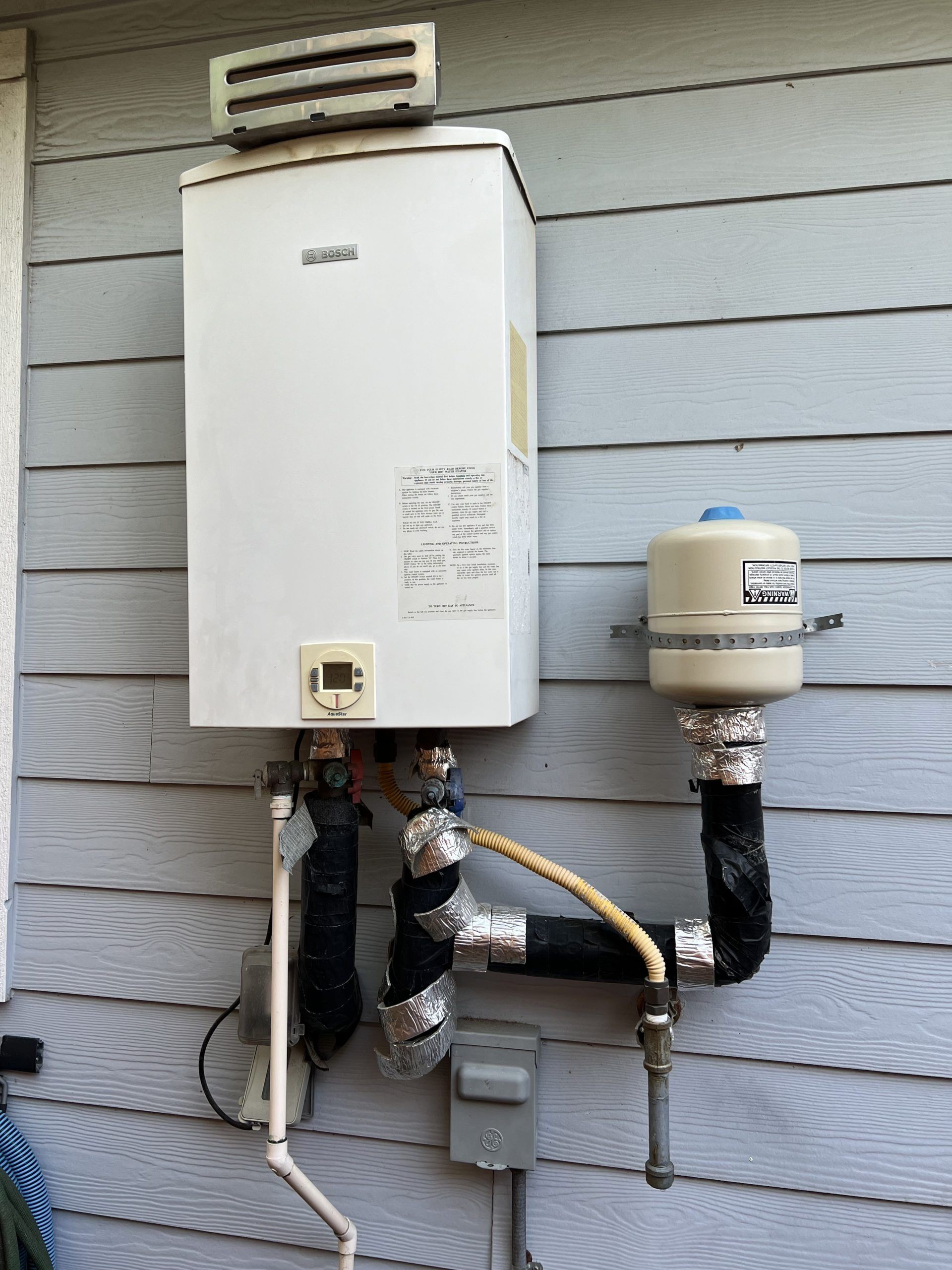 White tankless water heater mounted on a light gray wall; includes a storage tank, insulated pipes, and electrical box.